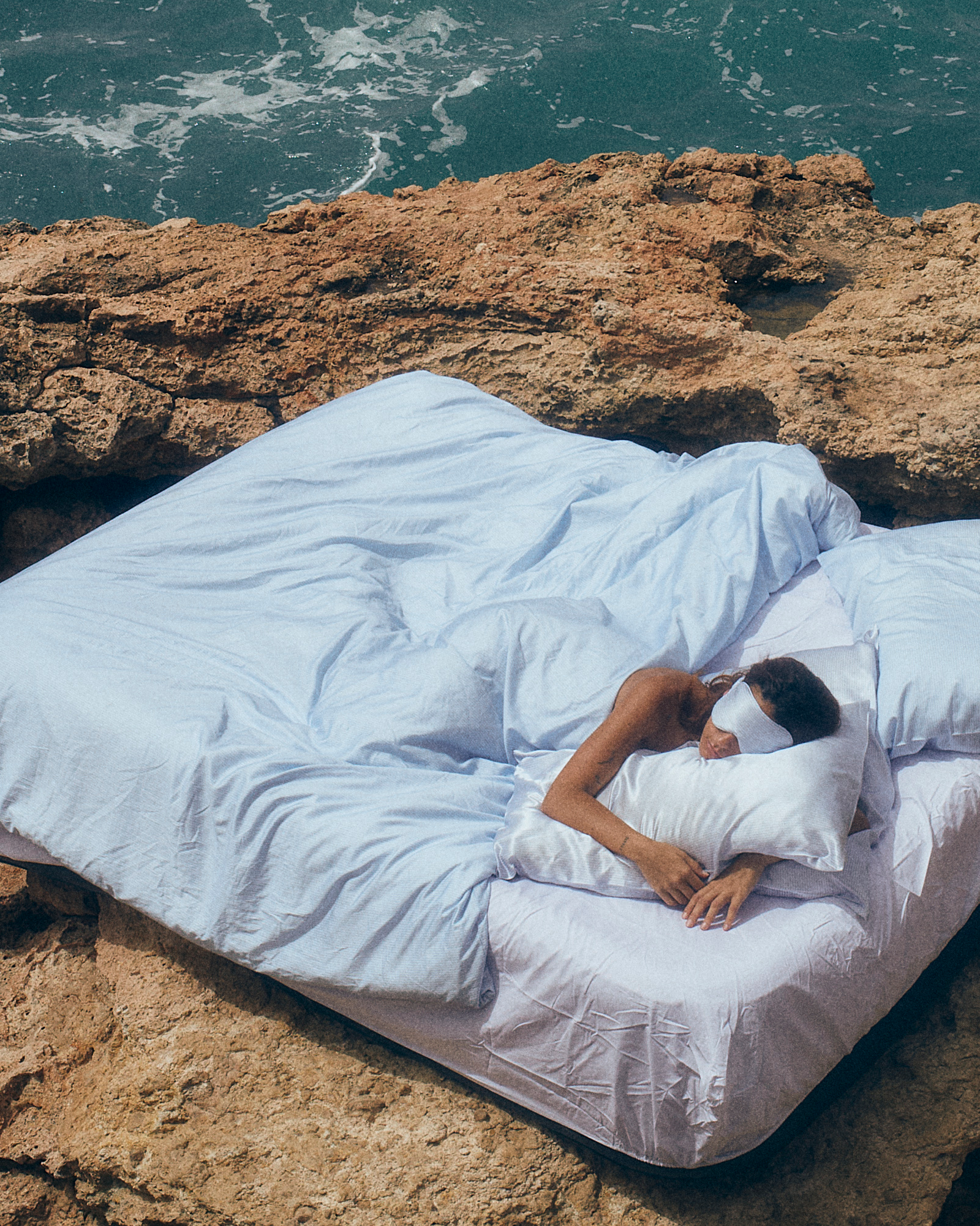 Person lying on a bed with white sheets and pillows on a rocky cliff overlooking the ocean.